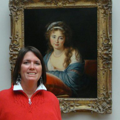 Julie Griffin standing in front of one of her masterpieces. Julie is wearing a red sweater and has dark brown hair. The painting behind her is a repruction of an original painting of a woman with a gold frame.